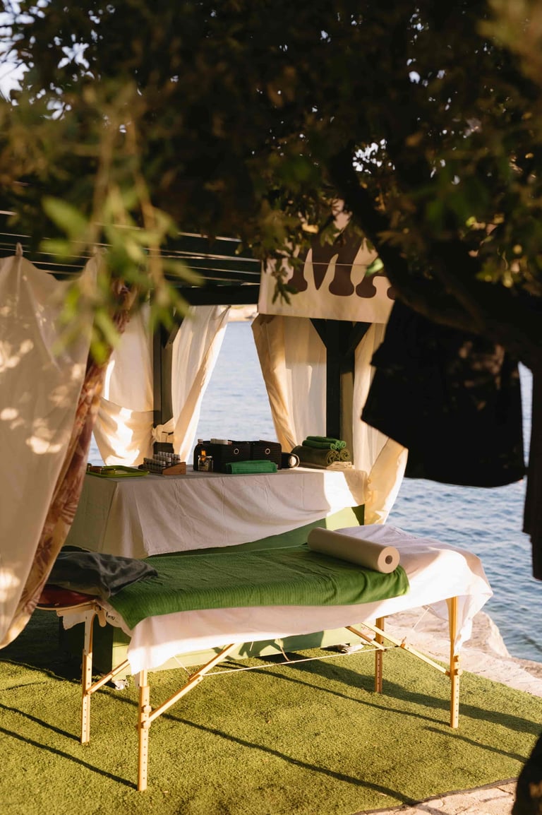 Outdoor spa setup with massage table under shaded pergola with flowing white fabric, overlooking water